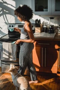 Woman in casual clothes and fabulous afro talks out of a kitchen area with a teacup in one hand, and a saucer in the other, with her mixed-breed terrier following at her feet