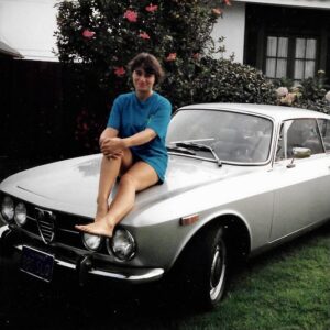 Teenaged Stacy sitting on the hood of a silver classic Alfa Romeo car, parked on the lawn of a house